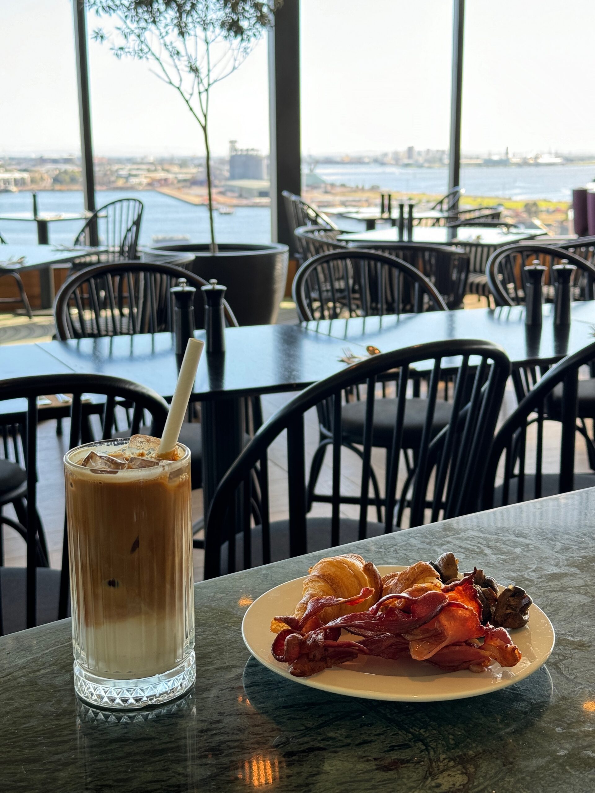 a plate of hot food breakky and a glass of iced oat milk latte on top of a table against a panoramic view of Newcastle Harbour, NSW