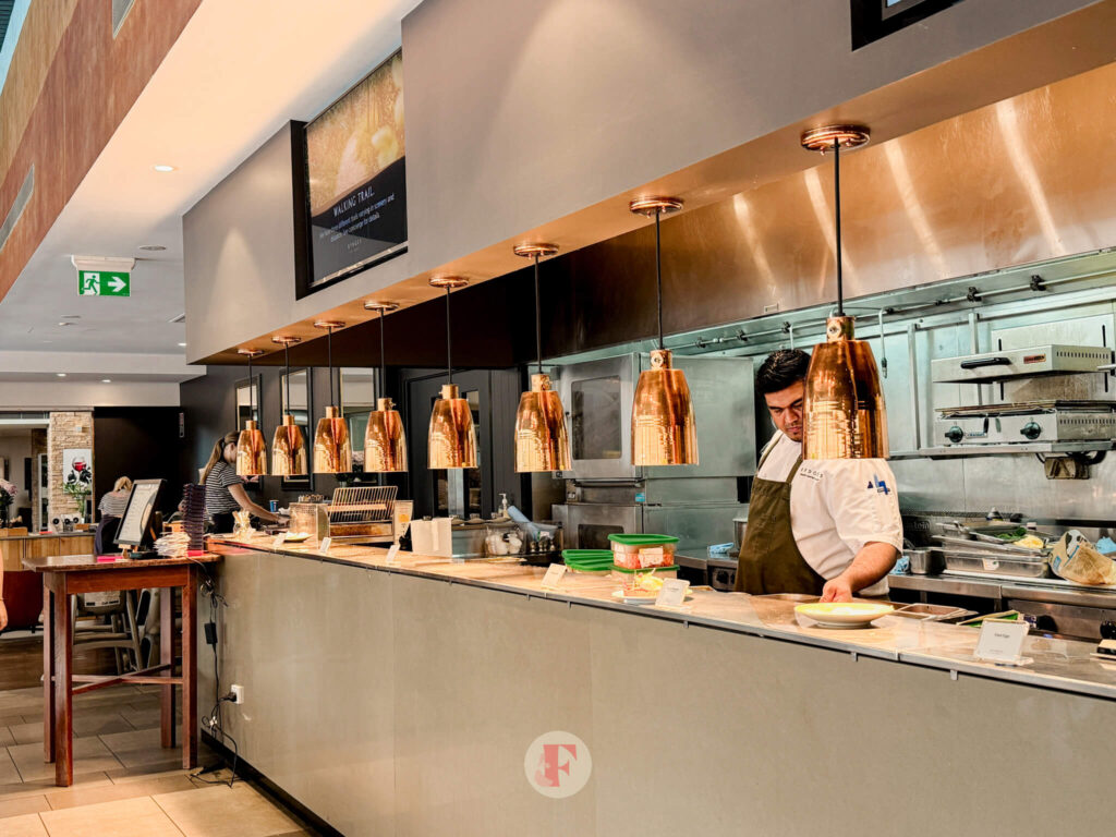 A chef preparing food at the hot food counter at the Rydges Hunter Valley breakfast buffet. Copper pendant lights hang above the counter, and the kitchen area is visible in the background.