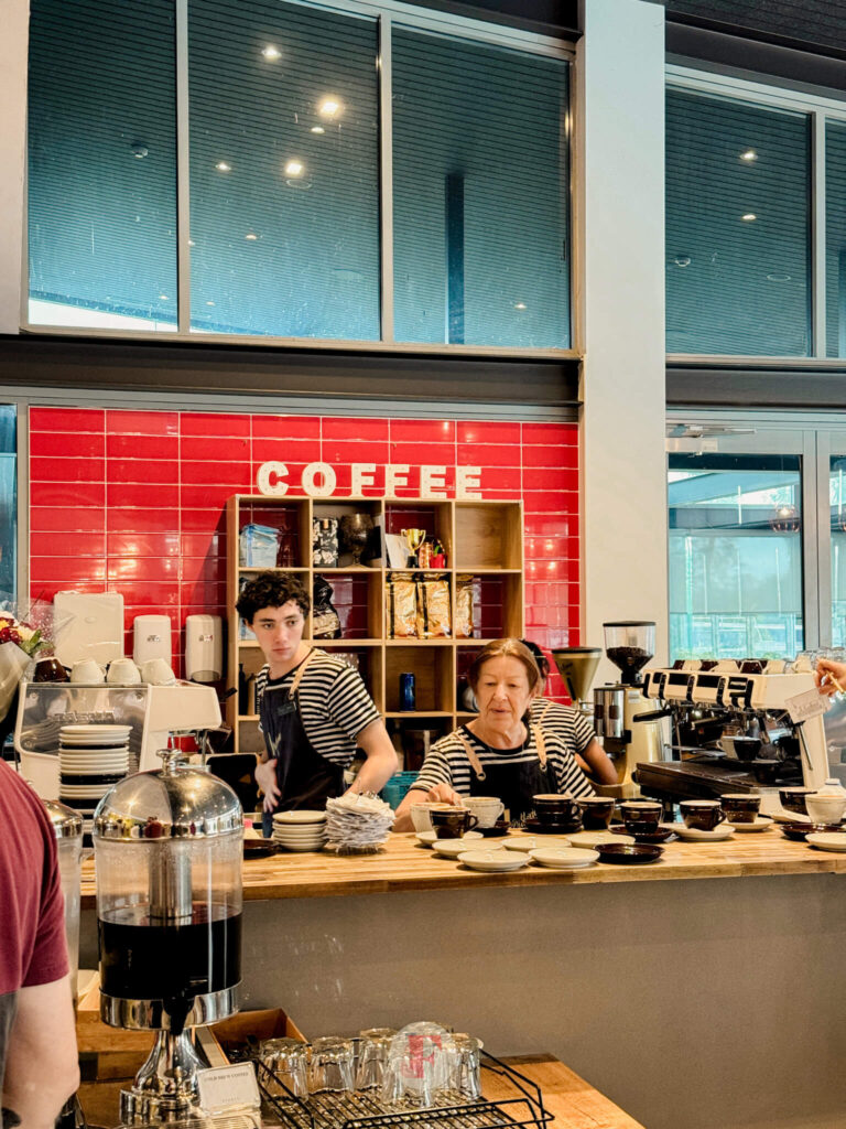 A coffee bar at Rydges Hunter Valley, with staff preparing coffee drinks. A bright red tiled wall in the background displays the word 'Coffee'.