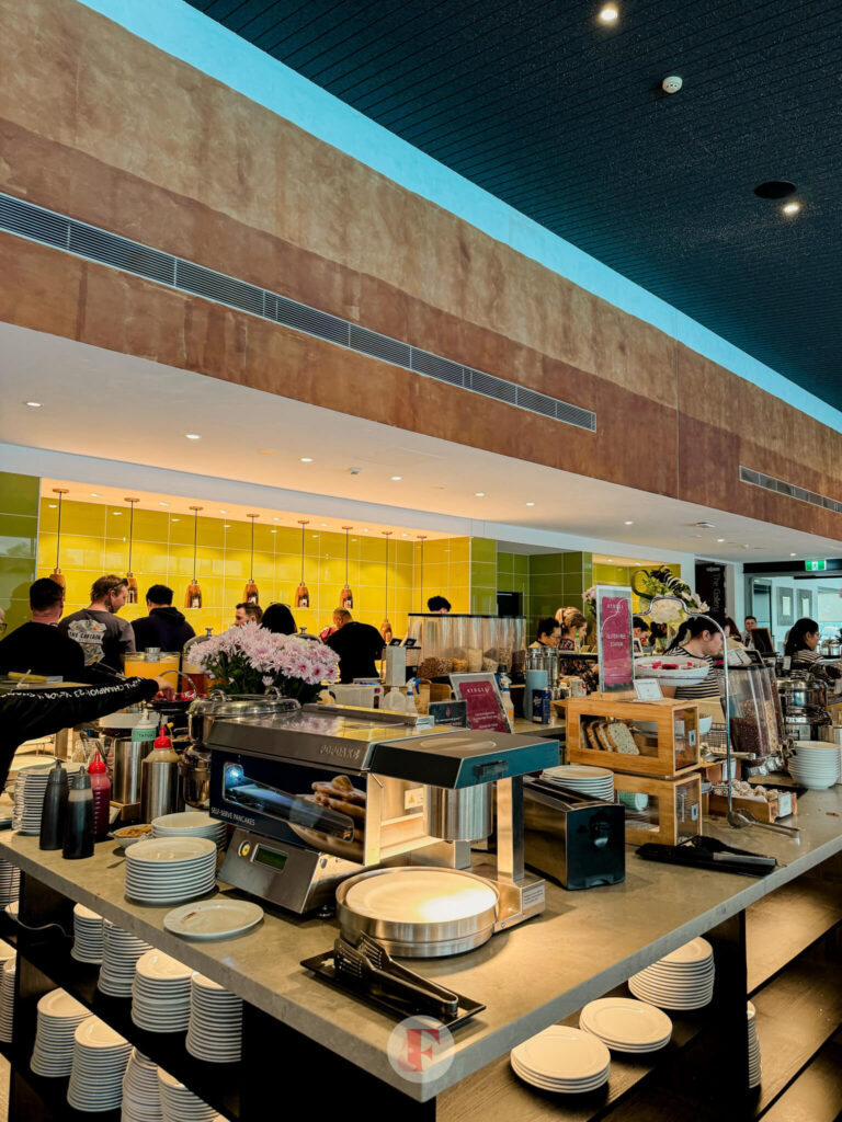 A section of the Rydges Hunter Valley breakfast buffet showing the pancake machine, plates, and various breakfast condiments, with a view of the kitchen area in the background.