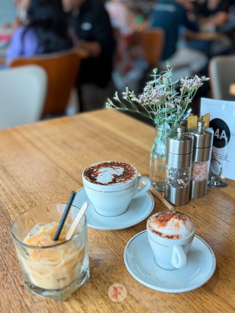 A table setting with cups of cappuccino, iced coffee, and a small flower arrangement, with a blurred background of other diners at the Rydges Hunter Valley breakfast buffet.