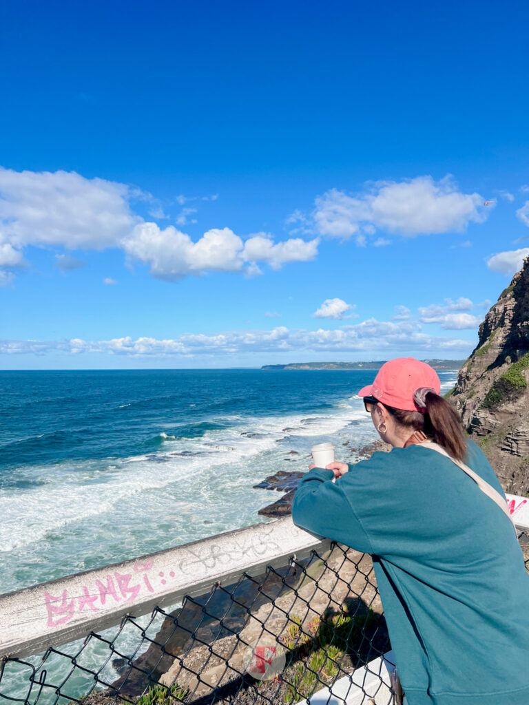A woman wearing green and an orange cap leaning onto a rail and staring at the ocean holding a cup of coffee