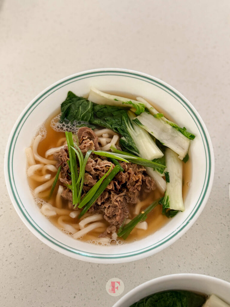 A top shot of a bowl of beef udon soup with slices of green onions and bok choy on top